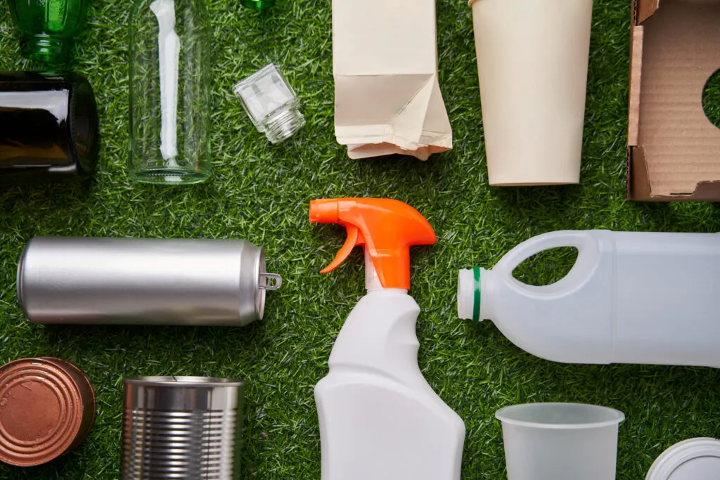 Różne rodzaje opakowań – butelki, puszki, kartony i plastikowe pojemniki ułożone na zielonym tle – ilustracja do sekcji opakowania i odpady opakowaniowe w kancelarii KRK